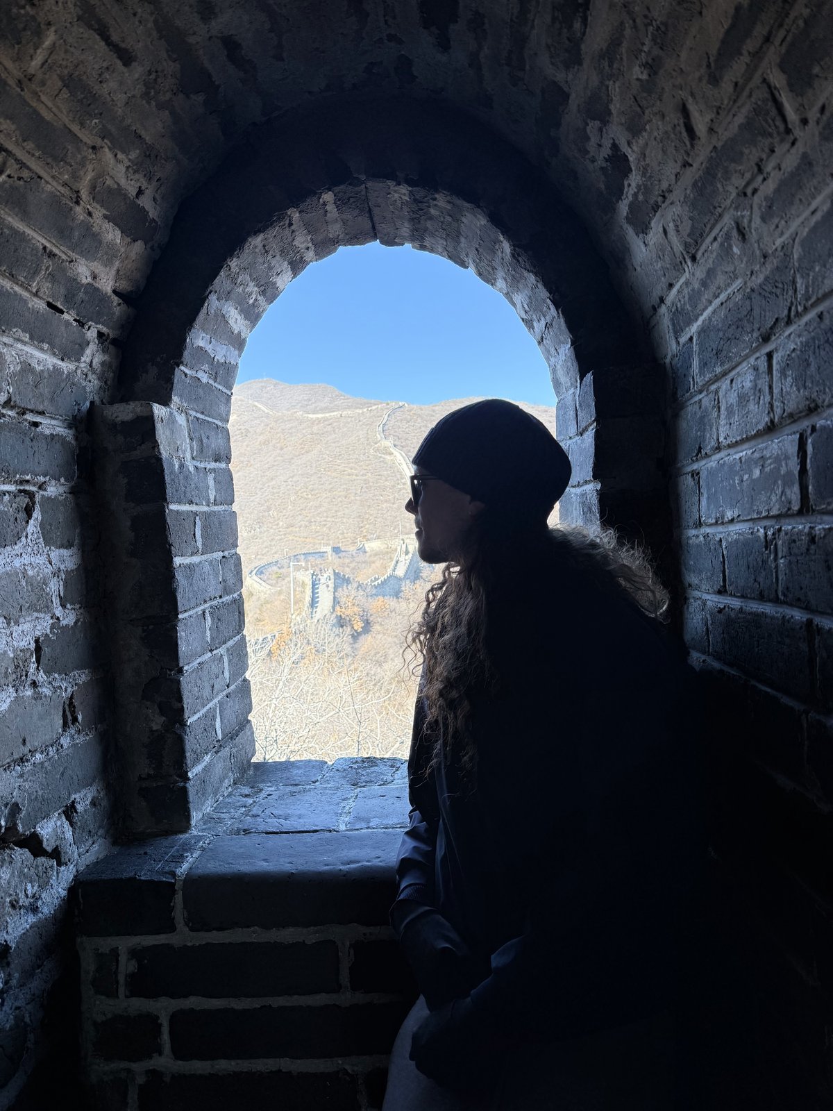 Hombre de cabello largo, gorro negro y gafas de sol, de perfil junto a una ventana de piedra. Al fondo, la Gran Muralla China recorre las montañas bajo un cielo azul.
